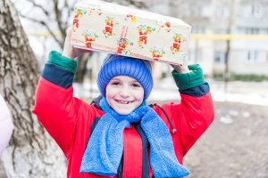 Smiling child in red coat and blue hat and scarf holding shoebox on head