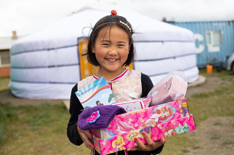 A YOUNG GIRL IN MONGOLIA SHOWS OFF THE TREASURES SHE RECEIVED IN HER SHOEBOX AND THE GOSPEL STORYBOOK IN HER LANGUAGE SHE WAS GIVEN ALONGSIDE IT.
