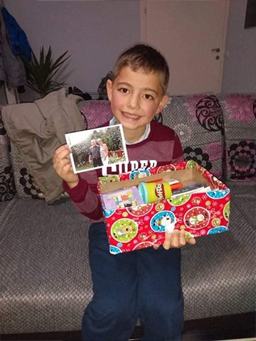 Boy with red wrapped shoebox smiles and holds photo from shoebox