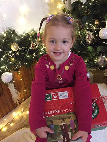 little girl with red shoebox smiles in front of Christmas tree